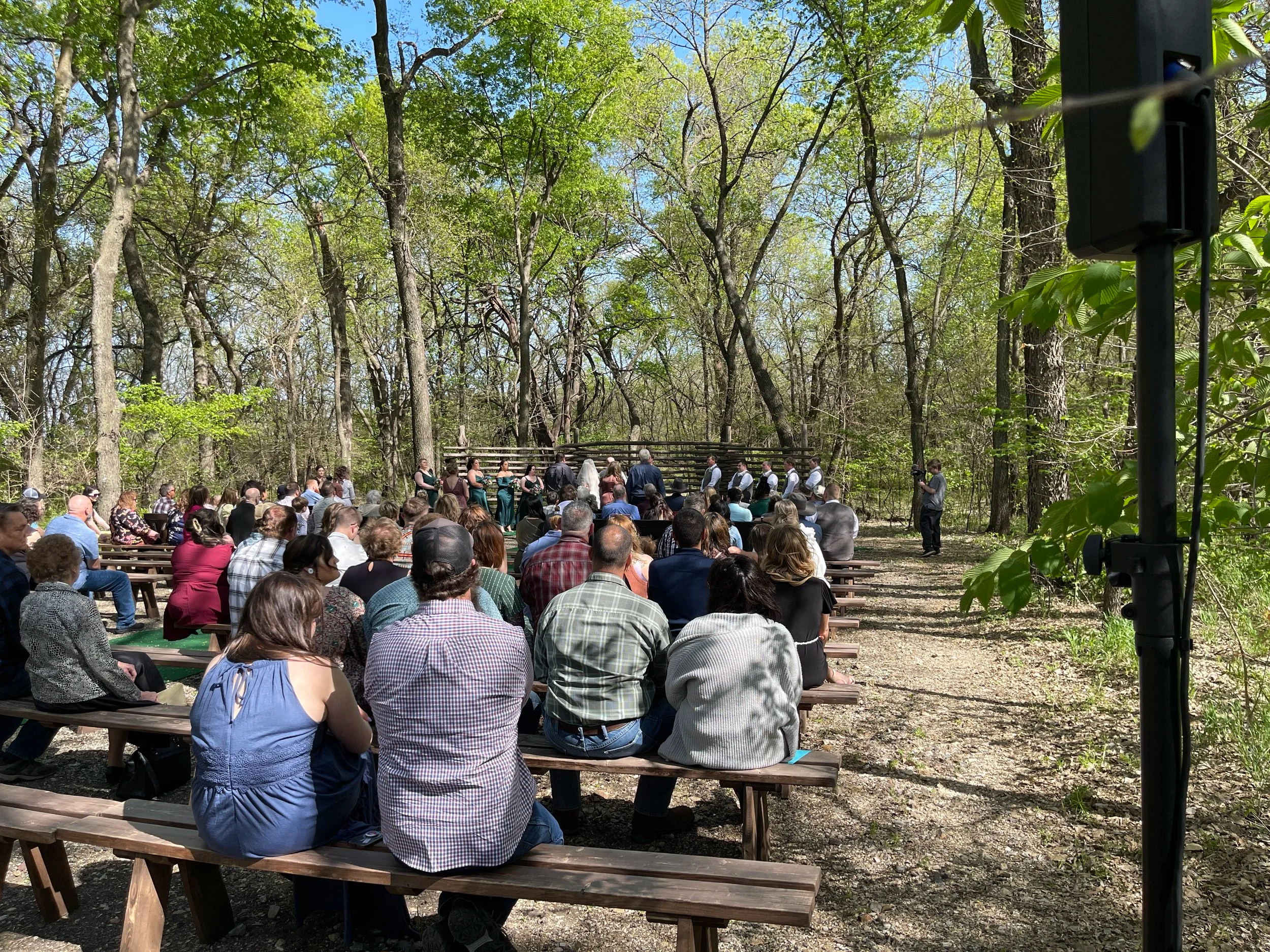 Guests on wooden benches at an outdoor wedding ceremony in the woods, with a speaker on a stand