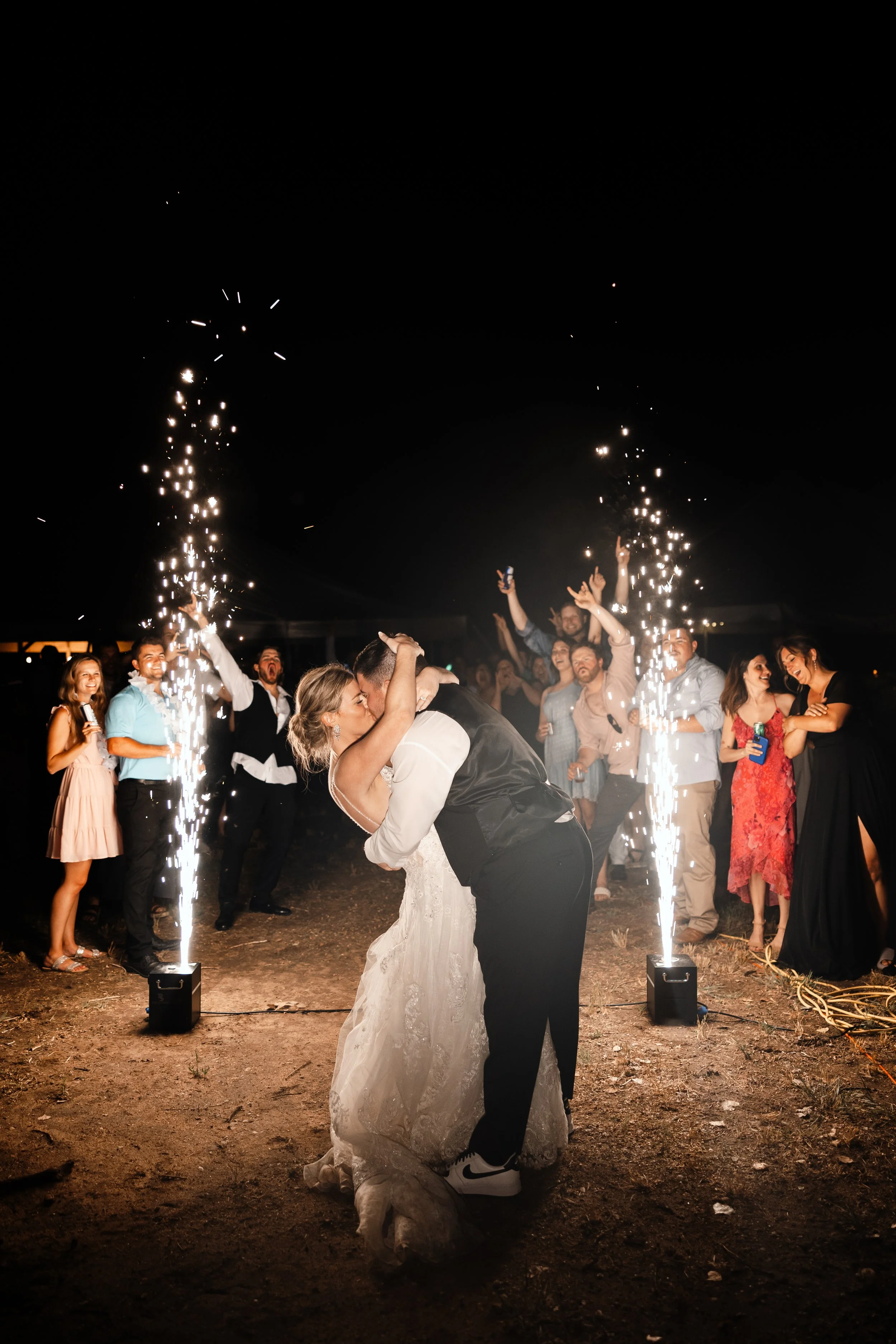 Newlyweds kiss between two cold spark fountains as guests cheer at night